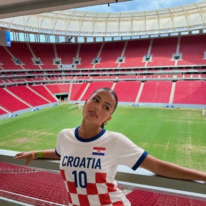 May include: White t-shirt with blue trim and the word "CROATIA" in blue. The shirt features a Croatian flag and the number 10. The bottom half of the shirt has a red and white checkered pattern. The background shows a stadium with red seating.