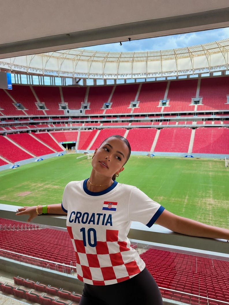 May include: White t-shirt with blue trim and the word "CROATIA" in blue. The shirt features a Croatian flag and the number 10. The bottom half of the shirt has a red and white checkered pattern. The background shows a stadium with red seating.