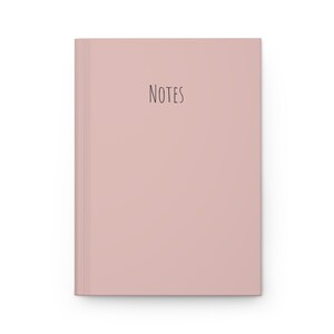 May include: A blush-colored notebook with the word "NOTES" in a simple, handwritten-style font. The notebook has a clean, minimalist design with a smooth cover and a rectangular shape.