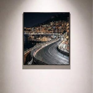 May include: A framed photograph of a nighttime cityscape with a winding road and a harbour filled with boats. City lights illuminate the buildings and water, creating a detailed scene. The image is displayed on a white wall, lit by a spotlight.