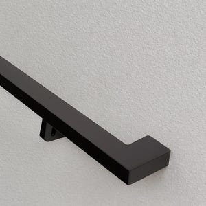 May include: A black, rectangular handrail is mounted on a textured, light gray wall. The handrail has a square profile and a right-angle turn at the end. The handrail is a modern design element.