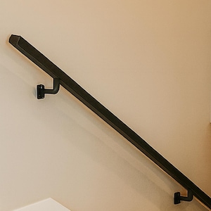 May include: A black metal handrail installed on a beige wall. The handrail is angled and supported by black brackets. The design is modern and minimalist, suitable for interior staircases or hallways.