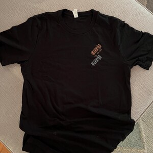 May include: Black crew neck t-shirt with a small graphic of two race cars in orange and blue. The text "Live the Ride" is above the cars. The shirt is made of a soft material.