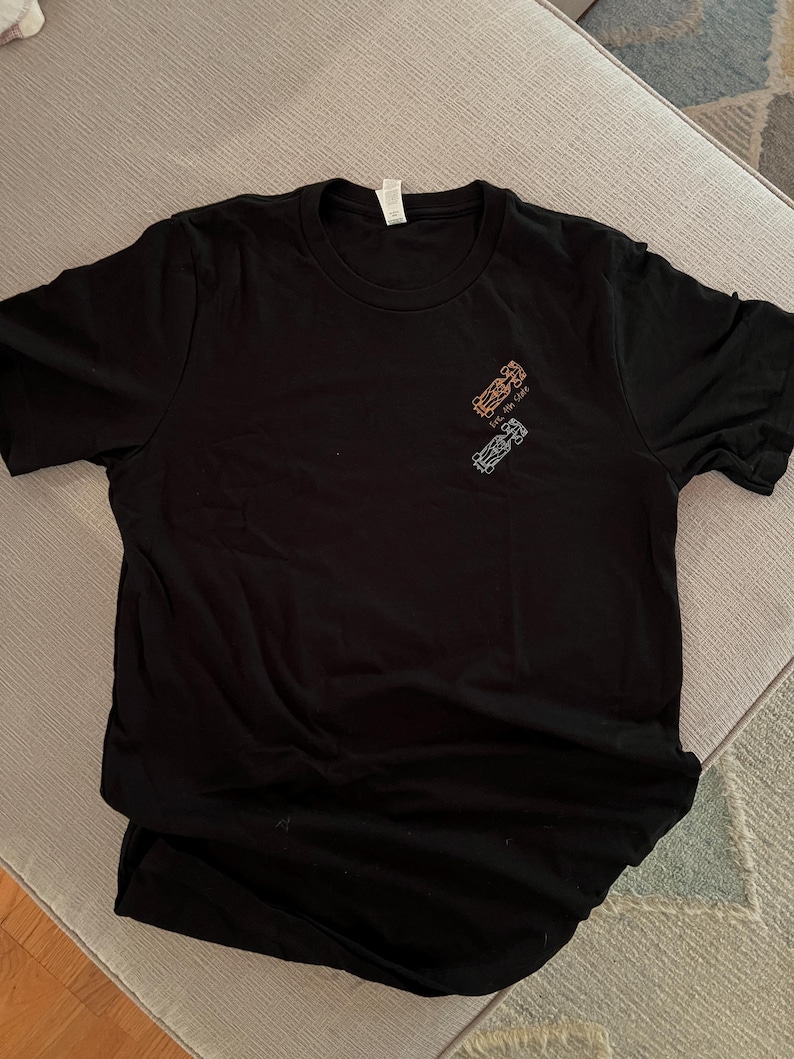 May include: Black crew neck t-shirt with a small graphic of two race cars in orange and blue. The text "Live the Ride" is above the cars. The shirt is made of a soft material.