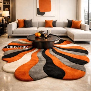 May include: A living room with a large sofa and orange cushions. A black coffee table displays pumpkins and candles. The room features a distinctive, flower-shaped rug in orange, black, grey, and cream, with the text "emee rug".