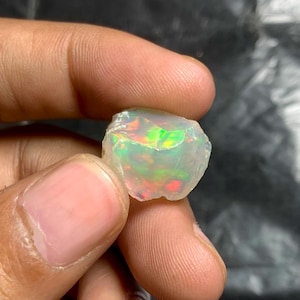May include: A raw, translucent opal gemstone displaying a vibrant play-of-color with flashes of neon green, orange, and red throughout its irregular, multifaceted surface. The stone has a milky, semi-transparent base with distinct internal light reflections.
