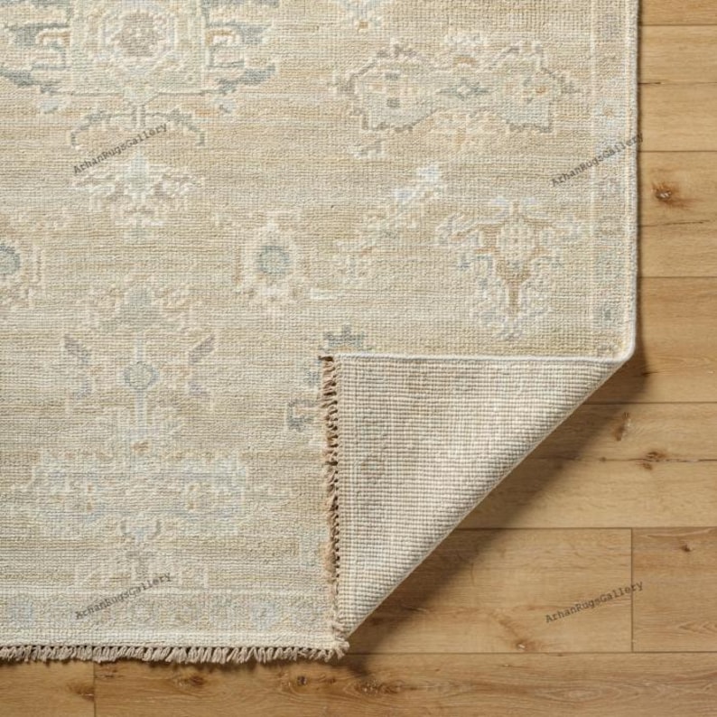 May include: Beige and light blue patterned area rug with a corner folded up, revealing the underside. The rug has a distressed, vintage look with a detailed floral design. The rug's edge has a fringe.