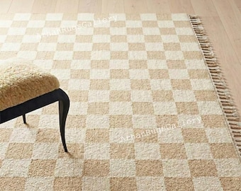Könnte beinhalten: Ein beige-cremefarbener karierter Teppich mit Fransen. Der Teppich hat ein Schachbrettmuster und besteht aus einem gewebten Material. Ein Teil eines Stuhls mit beigem Kissen und schwarzen Beinen ist sichtbar.