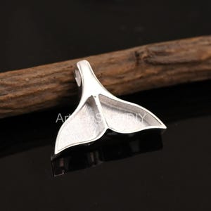 Può includere: Un ciondolo color argento a forma di coda di balena. Il ciondolo ha una superficie strutturata ed è posto su uno sfondo scuro con un elemento in legno. La coda è dettagliata con linee sottili.