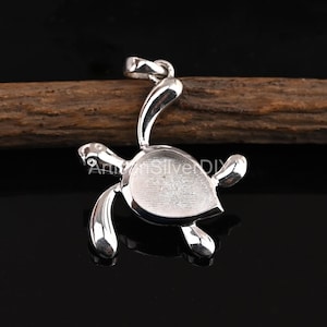 Puede incluir: Un colgante de tortuga plateada con un lazo para una cadena. La tortuga tiene un caparazón y aletas detalladas, y un cuello curvado. El colgante se muestra sobre un fondo oscuro.