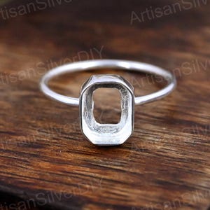 May include: A silver ring with an open rectangular setting. The ring's band is thin and delicate, and the setting is octagonal. The ring is displayed on a wooden surface.