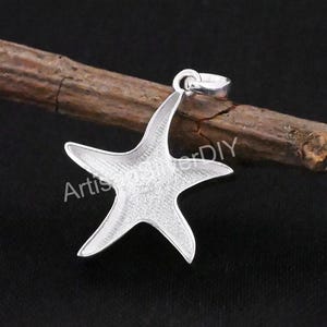Pode incluir: Pingente de estrela do mar prateado com superfície texturizada. O pingente tem uma argola na parte superior para uma corrente. A palavra "Artis" é visível no lado esquerdo do pingente, e "erDIY" no lado direito.