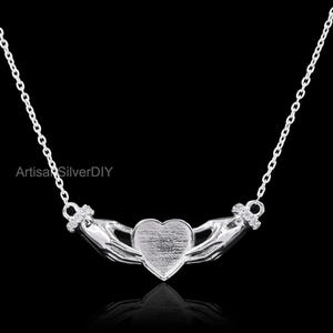 May include: A silver necklace with a heart-shaped pendant held by two hands. The heart has a textured finish, and the hands are intricately detailed. The chain is fine, and the design is set against a black backdrop.