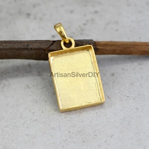 Puede incluir: Un colgante rectangular de color dorado con un lazo en la parte superior. El colgante tiene un área empotrada para engarzar una piedra u otro material. El texto "ArtisanSilverDIY" es visible en el colgante.