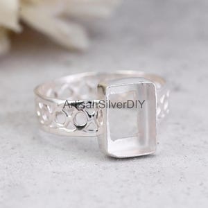 Puede incluir: Un anillo de plata con un engaste rectangular. La banda presenta un diseño calado decorativo. El anillo está sobre un fondo neutro. El texto "ArtisanSilverDIY" es visible en el anillo.