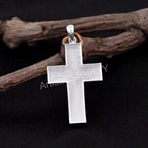 Könnte beinhalten: Ein silberner Kreuzanhänger mit strukturierter Oberfläche. Das Kreuz hängt an einer kleinen Schlaufe und einem braunen Ring. Der Hintergrund ist eine dunkle, verschwommene Oberfläche mit einem braunen Ast.