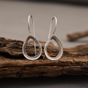 May include: A pair of silver teardrop earrings with a minimalist design. The earrings are displayed on a piece of dark wood. The text "ArtisanSilverDIY" is visible on the wood.