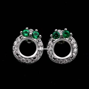 May include: A pair of silver-toned stud earrings. Each earring features a circular design with a ring of small, clear stones surrounding a central opening. Two green gemstones are set at the top of each earring.