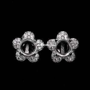 May include: A pair of silver-toned flower-shaped earring settings. Each setting features a central circular space surrounded by five rounded petals, each encrusted with small, sparkling stones. The earrings are set against a black background.