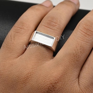 May include: A silver-colored rectangular ring with a recessed top, worn on a finger. The ring has a simple, modern design. The word "Artisan" is visible on the ring.