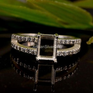 May include: A silver ring setting with a rectangular center and a split band. The band is adorned with small, clear stones. The ring is displayed on a reflective black surface, with the text "ArtisanSilverDIY" visible.