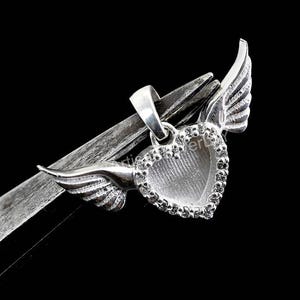 May include: A silver heart-shaped pendant with wings, set with small, clear stones. The pendant is held by a silver metal tool against a black backdrop. The wings are detailed and add a decorative element.