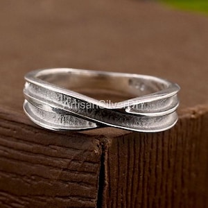 May include: A silver ring with a textured, interwoven design. The ring features a band that crosses over itself, creating a unique look. The ring is made of a shiny, metallic material.