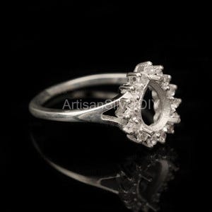 May include: A silver ring with a pear-shaped setting, encircled by small, sparkling stones. The ring's band is slender and polished, and the setting is designed to hold a gemstone. The ring is shown against a black background.