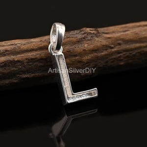 Pode incluir: Um pingente prateado em forma da letra "L" é exibido contra um fundo escuro. O pingente é feito de metal brilhante e tem uma argola na parte superior para uma corrente. O nome da loja Etsy "ArtisanSilverDIY" é visível.