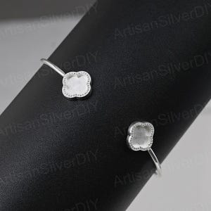 May include: A silver-colored cuff bracelet with two four-leaf clover shaped settings. The settings are outlined with small, sparkling stones. The bracelet is displayed on a black surface.