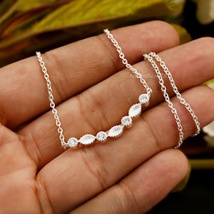 May include: A delicate silver necklace featuring a curved bar design. The bar is adorned with alternating round and marquise-shaped clear gemstones. The necklace is displayed on a hand, showcasing its fine chain and sparkling details.