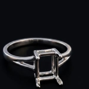 May include: A polished silver-toned metal ring setting featuring a rectangular four-prong head designed to hold a gemstone. The band has a split shank design that tapers as it connects to the central mounting, creating a clean and structural look.