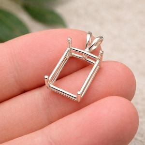 Puede incluir: Un engaste de colgante rectangular plateado con cuatro garras en cada esquina y un lazo para una cadena. El colgante se sostiene en una mano sobre un fondo borroso. El engaste está diseñado para sostener una piedra preciosa rectangular.