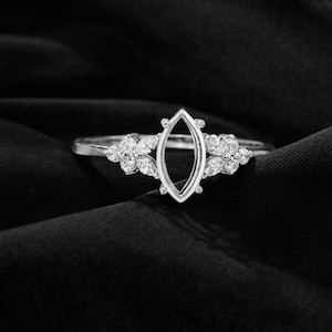 Peut inclure: Une bague en argent avec un sertissage central en forme de marquise, flanquée de grappes florales de petites pierres précieuses rondes. La bague est mise en valeur sur un fond noir, soulignant son design complexe et son éclat.