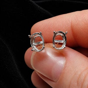 May include: Two silver oval earring settings are held in a person's hand. The settings have a cage-like design with four prongs at the corners to hold a gemstone. The background is black.