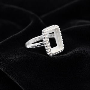 May include: A silver ring featuring a rectangular bezel setting with a beaded edge. The ring band is smooth and polished, leading to a central rectangular frame designed to hold a stone or insert, showcasing a metallic finish throughout.