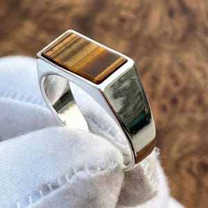 May include: A rectangular ring with a polished silver band and a rectangular tiger's eye stone. The tiger's eye stone has brown and gold bands. The ring is shown on a white surface.