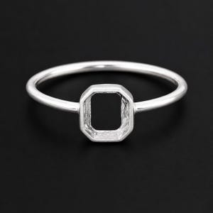 May include: A silver ring with an emerald-cut setting. The ring has a thin band and a square-shaped setting with beveled edges. The ring is set against a black background.