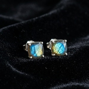 May include: A pair of square stud earrings with iridescent blue and yellow gemstones set in silver. The earrings are displayed on a black velvet surface, highlighting their vibrant colors and unique design. These earrings are a stylish accessory.