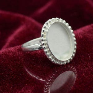 Peut inclure: Une bague en argent avec un sertissage ovale. Le sertissage est entouré de petites perles rondes. La bague est présentée sur une surface de velours rouge foncé. Le design de la bague est simple et élégant.