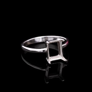 May include: A silver ring with a rectangular setting, designed to hold a gemstone. The ring is set against a black background, highlighting its reflective surface and intricate design. The ring's band is smooth and polished.