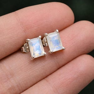Puede incluir: Un par de pendientes de botón rectangulares de piedra lunar. Los pendientes tienen un engaste plateado con cuatro pequeñas garras que sujetan la piedra lunar. Las piedras lunares tienen una base blanca lechosa con destellos azules y amarillos.