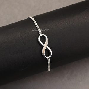 以下が含まれることがあります： シルバーのブレスレット。インフィニティシンボルのペンダントが特徴です。インフィニティシンボルが中心で、中央にテクスチャード加工のデザインがあります。ブレスレットは繊細なチェーンで、暗い背景に表示されています。「ArtisaSilverDIY」の文字が見えます。