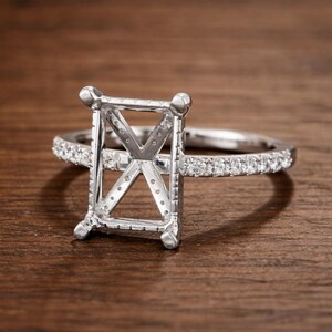 May include: A silver engagement ring setting with a rectangular center and a band lined with small, clear stones. The ring is set against a warm, wooden surface, highlighting its intricate design and sparkle.