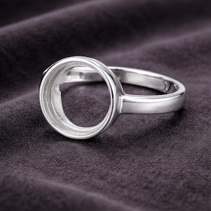 May include: A silver ring with a large, circular setting. The ring is made of polished metal and sits on a dark, textured surface. The setting is designed to hold a gemstone or other decorative element.