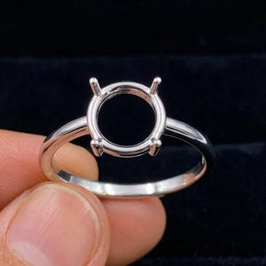 May include: A silver ring with a round setting designed to hold a gemstone. The ring has a simple band and four prongs to secure the stone. The ring is held by a hand against a dark background.