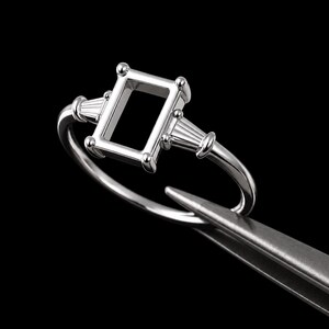 May include: A silver ring with an emerald-cut setting, flanked by two sets of three baguette-cut accents. The ring is held by a pair of tweezers against a black background, showcasing its intricate design and craftsmanship.