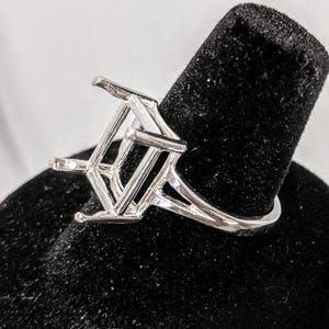 May include: A silver-colored ring with a geometric design. The ring features a square setting with angled supports, creating a modern and architectural look. The ring is displayed on a black velvet surface.