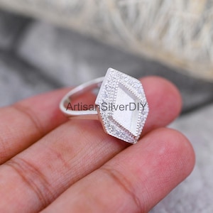 May include: A silver ring with a diamond-shaped setting. The setting is encrusted with small, clear stones around the edges and features a textured, rectangular center. The ring band is thin and polished.