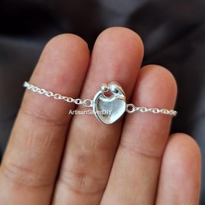 Può includere: Un braccialetto d'argento con un ciondolo a forma di cuore raffigurante due figure che si abbracciano. Il ciondolo è collegato a una catena delicata. Il testo "ArtisanSilverDIY" è visibile.